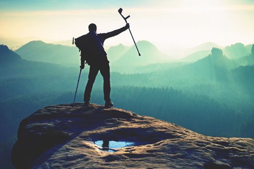 Hiker with medicine crutch above head achieved mountain peak. Broken leg fixed in immobilizer Deep misty valley bellow silhouette of man with hand in air. Spring daybreak