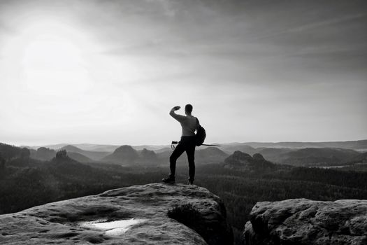 Tall backpacker with poles in hand. Sunny spring daybreak in rocky mountains. Hiker with big backpack stand on rocky view point above misty valley.