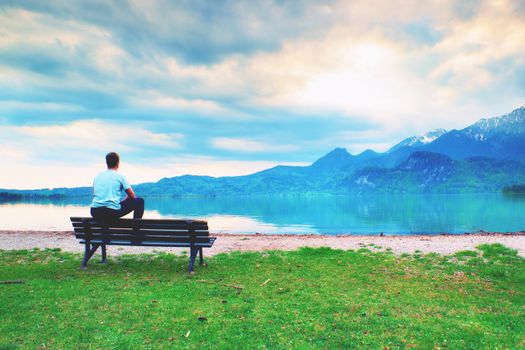 Tired adult man in blue shirt sit on old wooden bench at mountains lake coast. Vintage photo effect