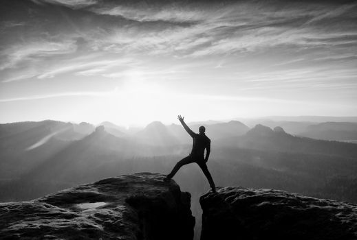 Jumping hiker in black gesture triumph between two rocky peaks. Wonderful daybreak in misty mountains.