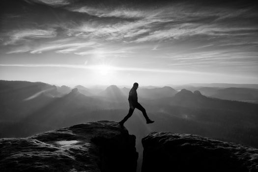 Crazy hiker in black is jumping between rocky peaks. Happy man. Wonderful daybreak in rocky mountains, heavy orange mist in deep valley. Miracle of nature