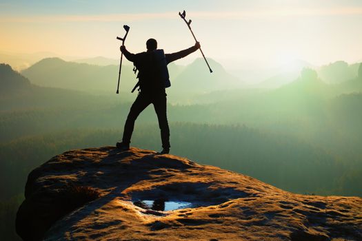 Tourist with  medicine crutch above head achieved mountain peak. Hiker with broken leg in immobilizer.  Deep misty valley bellow silhouette of man with hand in air. Spring daybreak