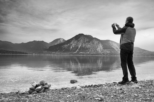 Tall man hold cellphone, take picture of autumn mountain lake scenery after rainy day. Vivid and vignetting effect. Poor lighting conditions.