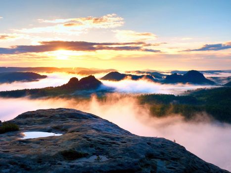 View over wet sandstone rocky peak into forest valley, daybreak Sun at horizon. Hills increased from foggy background
