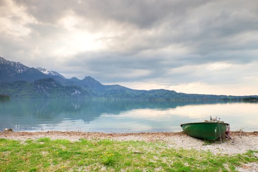 Abandoned fishing paddle boat on bank of Alps lake. Morning lake glowing by sunlight. Dramatic and picturesque scene. Mountains in water mirror.