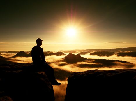 Moment of loneliness. Man with cap sit on the peak of rock and watching into colorful mist and fog in forest valley. Dreamy autumn ladscape