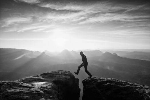 Crazy hiker in black is jumping between rocky peaks. Happy man. Wonderful daybreak in rocky mountains, heavy orange mist in deep valley. Miracle of nature