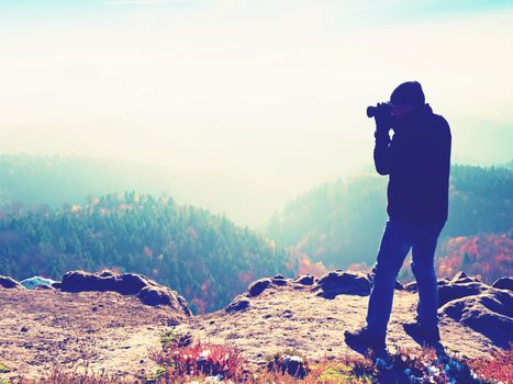 Photographer  on cliff. Nature photographer takes photos with mirror camera on peak of rock. Dreamy fogy landscape, spring orange pink misty sunrise in a beautiful valley below. 
