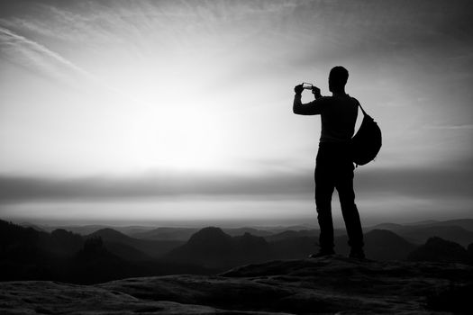 Tourist with backpack takes photos with smart phone on the rocky peak. Orange daybreak and fog in valley below