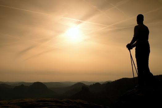 Silhouette of tourist with poles in hand. Sunny spring daybreak in rocky mountains. Hiker w on rocky view point above misty valley. 