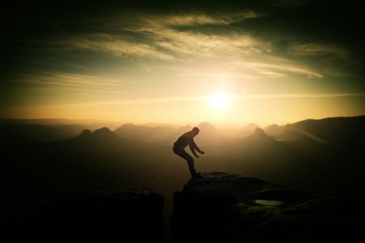 Crazy hiker umping between rocks. Marvelous colorful daybreak in rocky mountains, heavy orange mist in deep valley. Miracle of nature