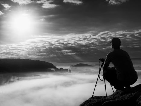 Amateur photographer takes photos with mirror camera on peak of rock. Dreamy fogy landscape, spring orange pink misty sunrise in a beautiful valley below