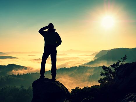 Man silhouette climbing high on cliff. Hiker climbed up to peak enjoy view. Man  watch over misty and foggy morning valley in morning Sun. 