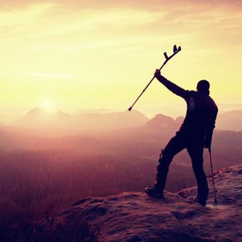 Hiker with  medicine crutch above head achieved mountain peak. Broken leg fixed in immobilizer Deep misty valley bellow silhouette of man with hand in air. Spring daybreak