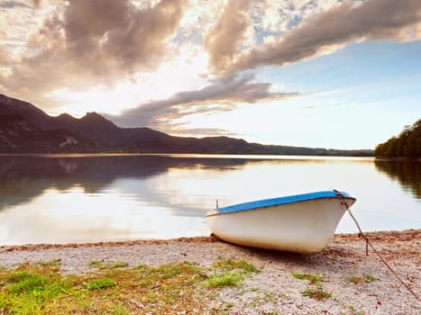 Abandoned white paddle boat on Alps lake bank. Evening lake glowing by sunlight. Dramatic and picturesque scene. Mountains in water mirror.