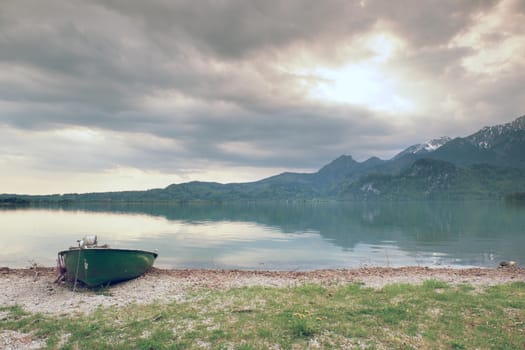 Abandoned fishing paddle boat on bank of Alps lake. Morning lake glowing by sunlight. Dramatic and picturesque scene. Mountains in water mirror.