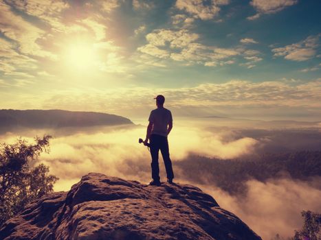 Tall photographer with baseball cap and tripod with camera in hands stand on rocky view point and watching down to deep misty valley bellow