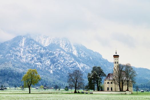 Old church with high bell tower and graveyard bellow mountains.  April weather. Wet spring snow in already fresh green grass  of meadow bellow mountains. 