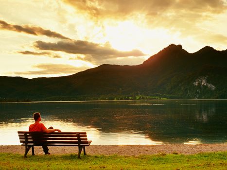 Alone man sits leg over on bench beside an azure mountain lake. Man relax and watch high peaks of Alps above lake mirror.