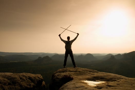 Happy hiker with x crossed poles above head.  Foggy  mountain valley bellow. Tall tourist enthusiast with poles in hands. Sunny spring daybreak in hills.