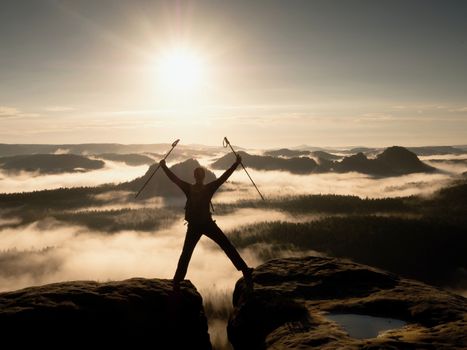 Hiker on peak. Tourist with bag in target with trekking poles in hand raised in air. Marvelous daybreak in fogy mountains