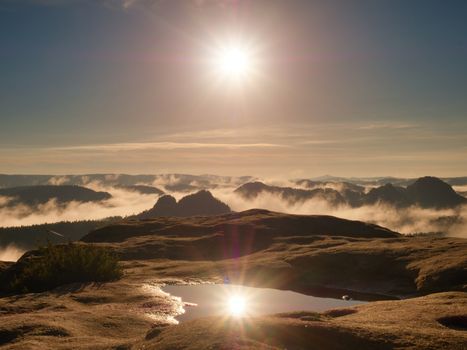 Water pool on cracked mountain summit.  Fairy daybreak in beautiful hills. Peaks of hills are in  yellow and orange fog stripped sun rays.