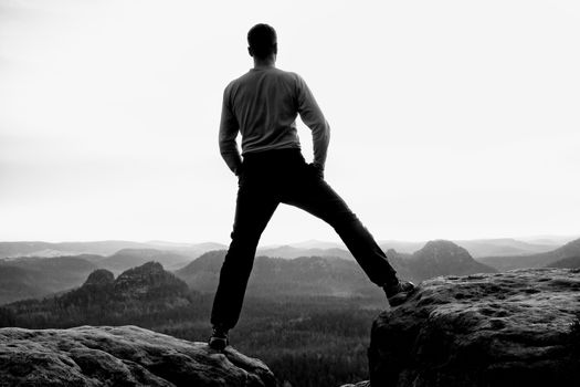 Happy man with raised arms gesture triumph on exposed cliff. Satisfy hiker silhouette on sandstone cliff watching down to hilly landscape. 