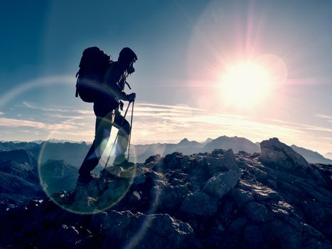 Lens flare light, strong defect. Tourist guide on trekking path  with poles and backpack.  Experienced hiker in windcheater and hood stand on rocky view point above misty valley. Sunny fall day