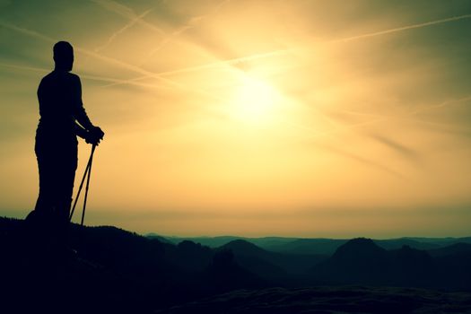 Silhouette of tourist with poles in hand. Sunny spring daybreak in rocky mountains. Hiker w on rocky view point above misty valley. 