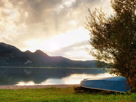 Upside down fishing paddle boat on bank of Alps lake. Smooth levelof autumnal lake. Dramatic and picturesque scene. Mountains in water mirror.
