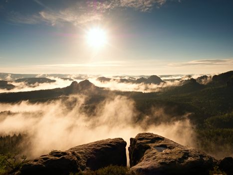Morning view over sandstone cliff into forest valley, daybreak Sun at horizon. Hills increased from foggy background