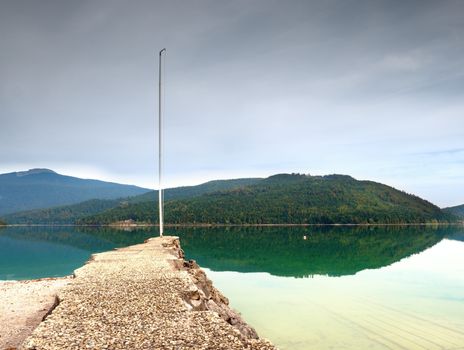 Stony sporty port at mountain lake. End of wharf with empty pole without flag. Dark blue clouds  in mirror of green blue water. Hills and mountain in backgorund.