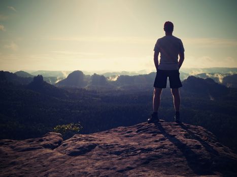 Young hiker in black pants and shirt on cliff edge and looking to misty hilly valley bellow. Enjoy life.Hiker sitting. Outdoor man enjoy.Cliff edge. Looking over horizon edge. Hike man.