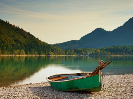 Abandoned fishing paddle boat on bank of Alps lake. Morning lake glowing by sunlight. Dramatic and picturesque scene. Mountains in water mirror.