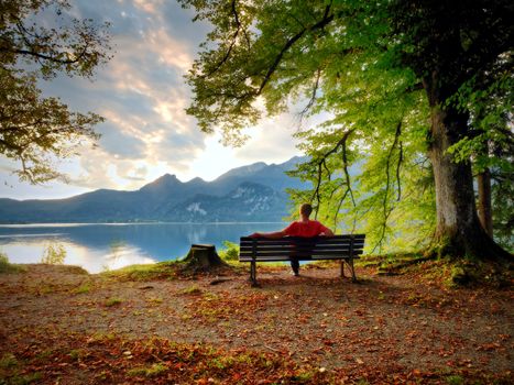 Man sit on wooden bench at mountain lake. Bank under beeches tree, mountains at horizon and in water mirror. Vintage toned photo.