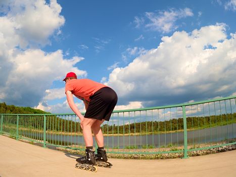 Rear view to inline skater in red t-shirt and black pants skating on the bridge . Outdoor inline skating on smooth concrete ground on lake bridge. Light skin man in four wheel boots