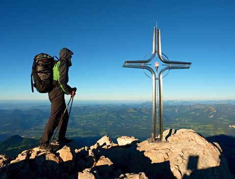 Hiker at big crucifix on mountain peak. Iron cross at Alps mountain top. Tourist with poles and heavy backpack  