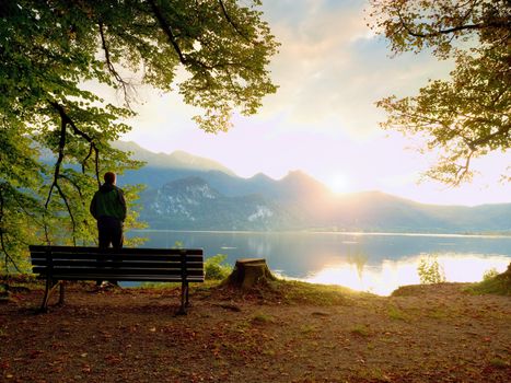 Man in green outdoor windcheater walk at lake bank.  Empty wooden bench, tree stump at mountain lake. Bank under beeches tree, mountains at horizon and in water mirror