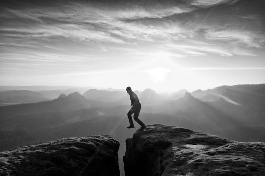 Man in black jumping between rocky empires. Dreamy daybreak in sandstone mountains, heavy orange mist in deep valley.