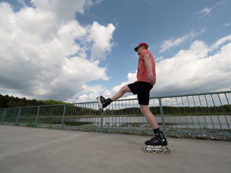 Middle age man in red t-shirt  with inline skates ride in summer park, popular outdoor roller skating. Sea bridge with blue sky in backgrround