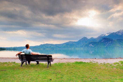 Alone  man sits on bench beside an azure mountain lake. Man relax and watch high peaks of Alps above lake mirror.