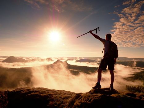 Tall backpacker with poles in hand. Sunny misty daybreak in rocky mountains. Hiker with backpack stand on rocky view point above valley. Vivid and strong vignetting effect.