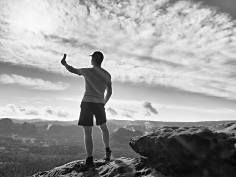 Man in tshirt and shorts takes photos with smart phone on peak of rock empire. Dreamy fogy landscape, spring orange pink misty sunrise in a beautiful valley of rocky mountains.