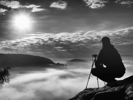 Amateur photographer takes photos with mirror camera on peak of rock. Dreamy fogy landscape, spring orange pink misty sunrise in a beautiful valley below