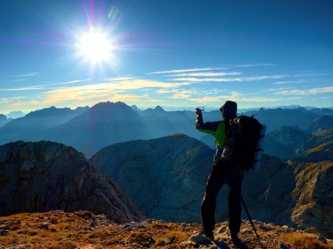 Experienced hiker takes phone photo. Man on Alps mountain peak.View to blue sky above deep foggy valley. Mountains increased from humidity