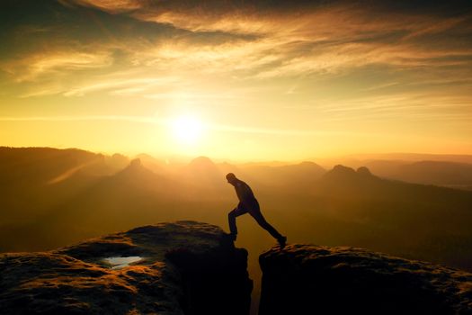 Crazy hiker in black is jumping between rocky peaks. Happy man. Wonderful daybreak in rocky mountains, heavy orange mist in deep valley. Miracle of nature