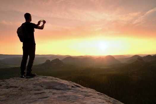 Tall man hiker is taking photo by smart phone on the peak of mountain at sunrise.