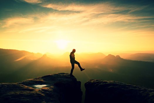 Man in black jumping between rocky empires. Dreamy daybreak in sandstone mountains, heavy orange mist in deep valley.