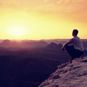 Adult tourist in black trousers, jacket sit on cliff's edge and looking to misty hilly valley bellow. Hikker relax and thinking alone in nature. 