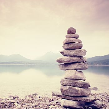 Balanced stone pyramide on shore of blue water of mountain lake. Blue mountains in water level mirror. Children built pyramid from pebbles.  Poor lighting conditions.